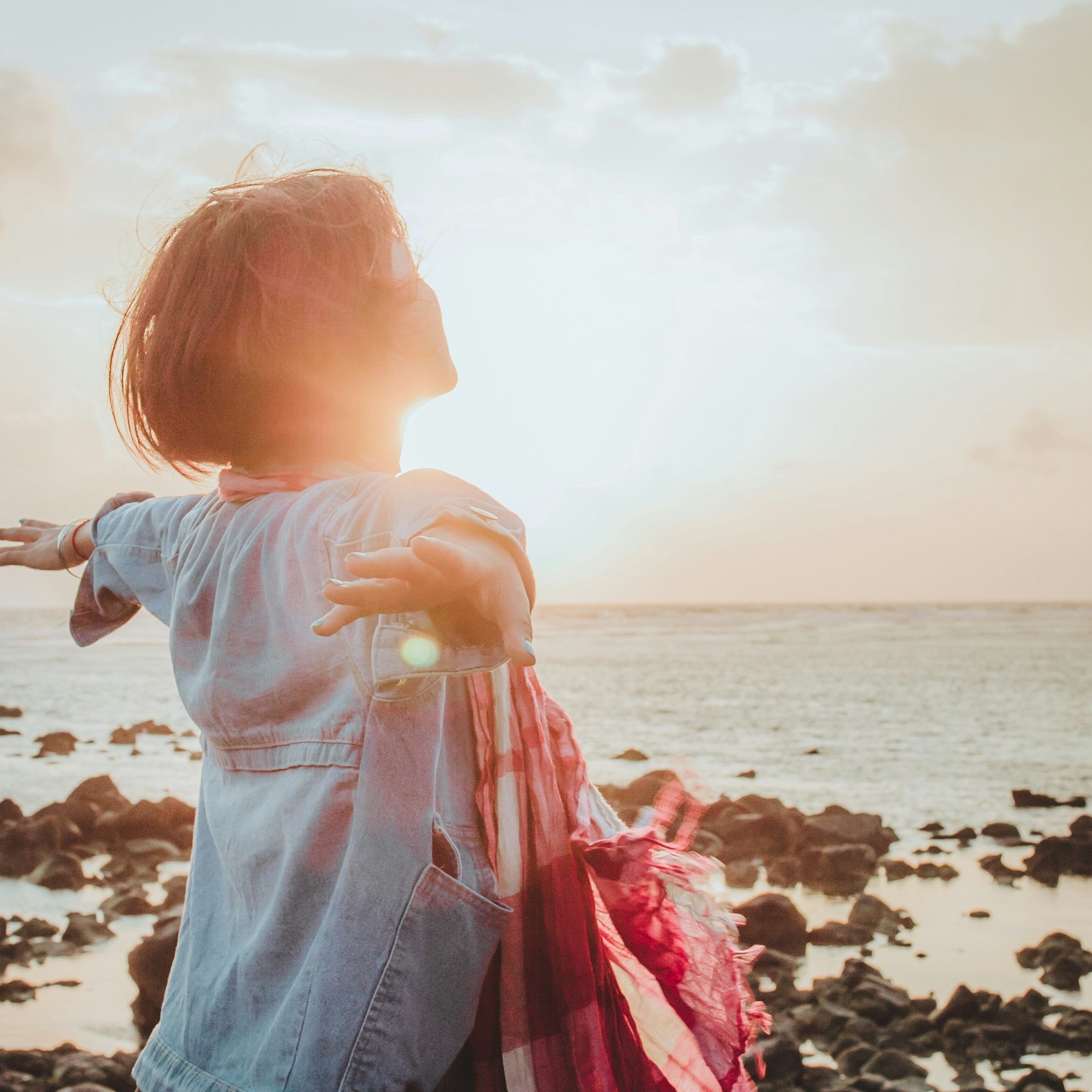 Woman standing on a rocky beach with the sun shining through, creating a warm glow.